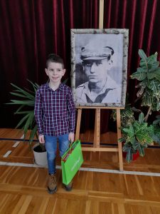 Wyróżniony w konkursie uczeń - Karol Cebulski pozuje do zdjęcia przy portrecie księdza Frelichowskiego. Chłopiec uśmiecha się a w prawej dłoni trzyma zieloną torbę z otrzymaną nagrodą.