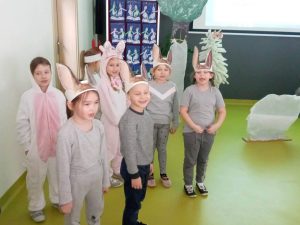 Uczniowie oddziału przedszkolnego podczas przedstawienia z okazji dnia babci i dziadka. Dzieci stoją w grupce. Na głowach mają opaski z długimi uszami. Część z nich szeroko otwiera buzię.