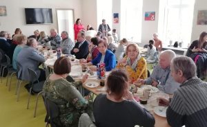 Babcie i dziadkowie uczniów oddziału przedszkolnego siedzą przy długich stołach ustawionych w dwóch rzędach na szkolnej stołówce. Na stołach stoją napoje i słodki poczęstunek.