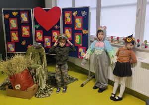 Troje uczniów oddziału przedszkolnego podczas przedstawienia z okazji dnia babci i dziadka. Dzieci stoją obok siebie. Dwoje z nich jest w przebraniach sowy a trzeci chłopiec jest w stroju wojskowego. Chłopiec ten podtrzymuje dłońmi czapkę. Obok niego ustawione jest krzeslo przykryte zionym kocem a przed nim worek ze słomą. Za dziećmi na rozstawionym tryptyku przyczepione są kolorowe kartki z kwiatami w czerwonych ramkach, wycięte z papieru filiżanki a po środku nich duże czerwone, papierowe serce.