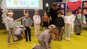 Uczniowie oddziału przedszkolnego wraz ze swoją wychowawczynią podczas wystąpienia z okazji dnia babci i dziadka. Dzieci stoją nierównomiernie rozstawione na sali teatralnej. Mają na sobie stroje przygotowane na występ. Dwoje uczniów kuca przy rozsypanych po podłodze kolorowych papierkach