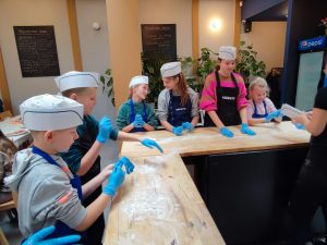 Uczniowie klasy czwartej i piątej podczas pracy na warsztatach pizzy. Dzieci stoją przy wysokim stole w kształcie litery L. Stół przed nimi został posypany mąką. Dzieci na głowie mają białe papierowe czapeczki a na dłoniach niebieskie lateksowe rękawiczkach.