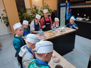 Uczniowie klasy czwartej i piątej podczas pracy na warsztatach pizzy. Dzieci stoją przy wysokim stole w kształcie litery L. Na stole przed nimi leżą rozwałkowane ciasto na pizze, na którym dzieci układają wybrane przez siebie dodatki wyłożone na stole w białych miseczkach. Uczniowie na głowach mają białe, papierowe czapeczki, a na dłoniach niebieskie, lateksowe rękawiczki.