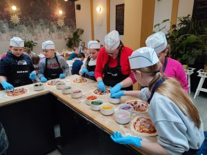 Uczniowie klasy piątej podczas warsztatów pieczenia pizzy. Dzieci stoją przy wysokim, drewnianym blacie. Na głowach mają białe, papierowe czapeczki, a na dłoniach niebieskie, lateksowe rękawiczki. Przed nimi na stole leżą przygotowywane pizze oraz miseczki z różnymi składnikami. Część dzieci wkłada dłonie do miseczek, inne umieszczają składniki na swoich pizzach.