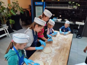 Uczennice w białych, papierowych czapeczkach oraz niebieskich, lateksowych rękawiczkach wałkują ciasto. Dziewczynki stoją przy wysokim drewnianym blacie posypanym mąką.
