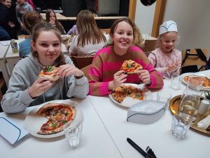 Trzy uczennice siedzą przy stole i jedzą przygotowane przez siebie pizze. Na stole przed nimi stoją talerze z pozostałymi kawałkami pizzy oraz szklanki do wody. W tle widać siedzących przy drugim stole innych uczestników wyjazdu.