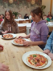 Dwie uczennice klasy czwartej stoją przy drewnianym blacie. Przed nimi na stole leżą na talerzach przygotowane przez nie pizze. Na stole leży jeszcze jedna pizza na białym talerzu. W tle widać siedzących przy długich stołach pozostałych uczestników wycieczki.