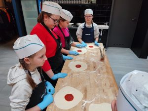Cztery uczennice klasy piątej stoją przy długim drewnianym blacie. Dziewczynki maja na głowach białe, papierowe czapeczki, a na dłoniach niebieskie, lateksowe rękawiczki. Na stole przed nimi leżą okrągłe placki pizzy z sosem pomidorowym umieszczonym po środku.