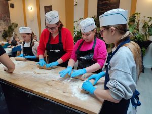 Uczennice klasy piątej stoją przy wysokim stole z drewnianym blatem i wałkują na nim ciasto na pizze. Dziewczynki mają na głowie białe, papierowe czapeczki, ubrane są w fartuchy z napisem azzurro, a na dłoniach mają niebieskie, lateksowe rękawiczki.