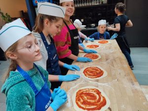 Uczennice z klasy czwartej i piątej stoją przy wysokim stole z drewnianym blatem. Dziewczynki mają założone na głowie białe, papierowe czapeczki. Przed nimi na stole leżą rozwałkowane i posmarowane przecierem pomidorowym placki pizzy.