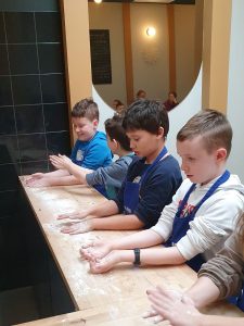 Uczniowie klasy czwartej stoją obok siebie przy wysokim stole z drewnianym blatem. Chłopcy rozsypują po stole mąkę.