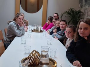 Uczniowie klasy piątej siedzą przy długim stole, na którym ustawiono karafki z wodą i szklanki.