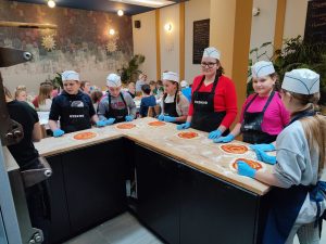 Uczniowie klasy piątej podczas pracy na warsztatach pizzy. Dzieci stoją przy wysokim stole w kształcie litery L. Na stole przed nimi leżą rozwałkowane ciasto na pizze posmarowane przecierem pomidorowym. Dzieci na głowie mają białe papierowe czapeczki a dłonie w niebieskich lateksowych rękawiczkach opierają o blat stołu. W tle widać siedzących przy długich stołach innych uczniów.