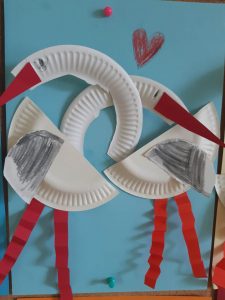 Zdjęcie pracy plastycznej wykonanej z okazji walentynek. Praca wykonana metodą wyklejania. Na niebieskiej kartce papieru wyklejono z kawałków tektury dwa łabędzie , których szyje nachodzą na siebie.