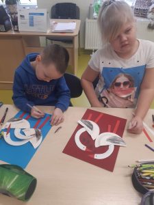 Dwoje uczniów oddziału przedszkolnego podczas wykonywania swoich prac plastycznych. Dzieci siedzą przy stoliku. Przed nimi leżą kartki z wyklejonymi łabędziami. Chłopiec trzyma w dłoni długopis a dziewczynka kawałek papieru.