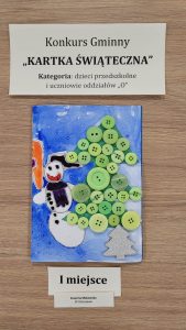 Praca plastyczna nagrodzona w konkursie "Kartka Świąteczna". Nad nagrodzona kartką prostokątna karteczka z nadrukiem :"Konkurs Gminny "KARTKA ŚWIĄTECZNA" Kategoria: dzieci przedszkolne i uczniowie oddziałów "0"". Pod nagrodzona kartka karteczka z nadrukiem "I miejsce" i jeszcze jedna mniejsza z nadrukiem "Zuzanna Makowska SP Ostaszewo". Kartka przedstawia bałwanka stojącego obok choinki wyklejonej zielonymi guzikami.