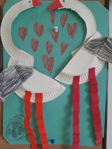 Zdjęcie pracy plastycznej wykonanej z okazji walentynek. Praca wykonana metodą wyklejania. Na zielonej kartce papieru wyklejono z kawałków tektury dwa łabędzie , które zbliżają do siebie dzioby. Pomiędzy ptakami narysowane są czerwone serduszka.