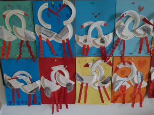 Zdjęcie ośmiu prac plastycznych przyczepionych do tablicy. Każda praca jest wykonana na kartce innego koloru i przedstawiaj dwa łabędzie obejmujące się.