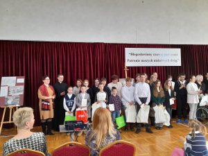 Zdjęcie grupowe nagrodzonych uczestników konkursu stojący wraz z jego organizatorami. Wszyscy stoją na tle czerwonej kurtyny na której widnieje baner z napisem "Błogosławiony czuwaj nad nami Patronie naszych niełatwych dróg".