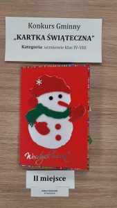 Praca plastyczna nagrodzona w konkursie "Kartka Świąteczna". Nad nagrodzona kartką prostokątna karteczka z nadrukiem :"Konkurs Gminny "KARTKA ŚWIĄTECZNA" Kategoria: uczniowie klas IV-VIII". Pod nagrodzona kartka karteczka z nadrukiem "I I miejsce" i jeszcze jedna mniejsza z nadrukiem "Robert Czachowski SP Ostaszewo". Kartka przedstawia bałwanka ubranego w czerwoną czapeczkę i rękawiczki oraz zielony szalik. Pod nim złoty napis Wesołych Świąt.
