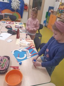 Dwie uczennice oddziału przedszkolnego stoją przy długim stole, na którym rozłożone są przybory plastyczne. Przed dziewczynkami leżą ich prace plastyczne przedstawiające łabędzie. Uczennica stojąca na pierwszym planie trzyma w dłoniach klej.