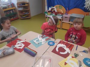 Trzy uczennice oddziału przedszkolnego siedzą przy wspólnym stole. Dwie z dziewczynek patrzą na siebie. Na stole leżą przybory plastyczne i prace, które wykonują uczennice.