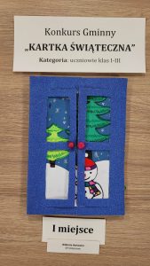 Praca plastyczna nagrodzona w konkursie "Kartka Świąteczna". Nad nagrodzona kartką prostokątna karteczka z nadrukiem :"Konkurs Gminny "KARTKA ŚWIĄTECZNA" Kategoria: uczniowie klas I-III". Pod nagrodzona kartka karteczka z nadrukiem "I miejsce" i jeszcze jedna mniejsza z nadrukiem "Wiktoria Hartowicz SP Ostaszewo". Kartka przedstawia bałwanka stojącego na tle białych górek z choinkami. Rysunek w tle zamkniętych niebieskich okiennic.