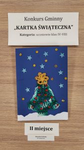 Praca plastyczna nagrodzona w konkursie "Kartka Świąteczna". Nad nagrodzona kartką prostokątna karteczka z nadrukiem :"Konkurs Gminny "KARTKA ŚWIĄTECZNA" Kategoria: uczniowie klas IV-VIII". Pod nagrodzona kartka karteczka z nadrukiem "I I miejsce" i jeszcze jedna mniejsza z nadrukiem "Weronika Zielińska SP Ostaszewo". Kartka przedstawia choinkę wykonaną z włóczki na niebieskim tle ozdobionym gwiazdkami i złotymi kółeczkami.