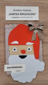 Praca plastyczna nagrodzona w konkursie "Kartka Świąteczna". Nad nagrodzona kartką prostokątna karteczka z nadrukiem :"Konkurs Gminny "KARTKA ŚWIĄTECZNA" Kategoria: uczniowie klas I-III". Pod nagrodzona kartka karteczka z nadrukiem "wyróżnienie" i jeszcze jedna mniejsza z nadrukiem "Lena Stalmierska SP Ostaszewo". Kartka przedstawia głowę Świętego Mikołaja naklejoną na kartkę i ozdobioną w prawym rogu kokardą.