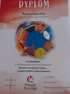 Zdjęcie dyplomu otrzymanego w konkursie przez Martynę Gajewską. Na czerwono białym tle lekko rozmazanym napisy: "Dyplom dla Martyny Gajewskiej ze Szkoły Podstawowej w Ostaszewie". Poniżej grafika w kształcie koła na którym umieszczono budynki na niebieskim tle. Pod grafiką napisy: "za zwycięstwo w XIV Powiatowym Konkursie Plastycznym "Mieszkam w ciekawym miejscu...podążam Szlakiem Kopernikowskim" . Poniżej podpis starosty Toruńskiego. Obok logo powiatu Toruńskiej oraz skład jury.