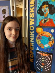 Nagrodzona w konkursie uczennica Martyna Gajewska pozuje do zdjęcia na tle galerii stworzonej z nagrodzonych prac plastycznych.