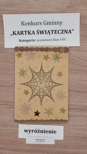Praca plastyczna nagrodzona w konkursie "Kartka Świąteczna". Nad nagrodzona kartką prostokątna karteczka z nadrukiem :"Konkurs Gminny "KARTKA ŚWIĄTECZNA" Kategoria: uczniowie klas I-III". Pod nagrodzona kartka karteczka z nadrukiem "wyróżnienie" i jeszcze jedna mniejsza z nadrukiem "Zofia Gajewska SP Ostaszewo". Kartka przedstawia Geometryczna gwiazdę wykonana cienkim czarnym długopisem w otoczeniu innych złotych gwiazdek. Całość wykonana na brązowym papierze.