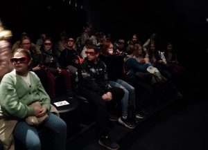 Uczniowie klasy piątej siedzą w ciemnej sali w rzędach. Uczniowie mają na oczach okulary 3D i rozglądają się po bokach.