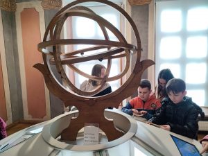 Zdjęcie eksponatu w muzeum. Wykonana z drewna konstrukcja złożona jest z umieszczonych jeden w drugim okręgu. Czworo uczniów stoi za konstrukcją i spogląda na monitor zamontowany pod nią.