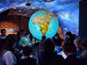 Uczniowie klasy piątej podczas zwiedzania muzeum. Uczniowie zgromadzeni są wokół podświetlonego, kolorowego globusa zamontowanego na suficie pomalowanym na niebiesko ze świecącymi się białymi kropkami imitującymi nocne niebo. Uczniowie pochylają się nad monitorami umieszczonymi w okręgu pod globusem. Jeden z uczniów wyciąga rękę w próbie dotknięcia świecącej kuli.