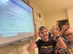 Uczniowie klasy drugiej stoją przy tablicy interaktywnej. Chłopcy podnoszą w górę ręce z wyprostowanymi dwoma palcami. Chłopcy uśmiechają się.