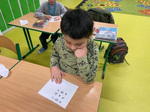 Uczeń klasy drugiej siedzi przy stoliku. Opiera dłoń na białej kartce z nadrukowanymi czarnymi rysunkami szyfru. Chłopiec pochyla się nad kartką. W tle widać innego ucznia siedzącego przy stoliku.