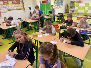 Uczniowie klasy drugiej siedzą przy stolikach w swojej sali. Przed dziećmi leżą kartki papieru. Część z nich pochyla się nad kartkami z długopisem w dłoni.
