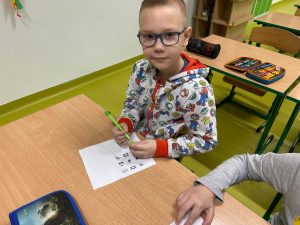 Uczeń klasy drugiej siedzi przy stoliku. Opiera dłoń na białej kartce z nadrukowanymi czarnymi rysunkami szyfru. Chłopiec chłopiec trzyma w dłoniach długopis. Patrzy przed siebie.