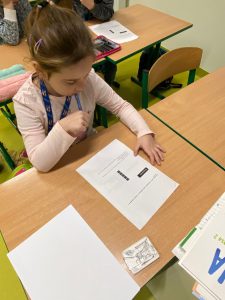 Uczennica klasy drugiej siedzi przy stoliku. Przed nią leży kartka papieru z nadrukowanymi zadaniami. Dziewczynka trzyma w dłoni długopis i patrzy na kartkę.