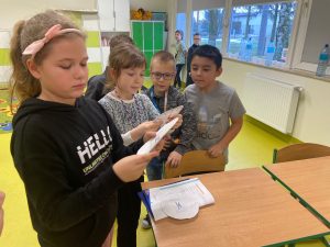 Uczniowie klasy drugiej stoją w małej grupce. Dwie uczennice stojące z przodu trzymają w dłoniach wycięte z papieru koła i przyglądają się im.