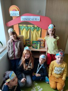 Uczniowie pozują do wspólnego zdjęcia przy stoliku teatrzykowym na którym umieszczono napis "DZIEŃ DINOZAURA". Dzieci mają na głowie papierowe opaski.