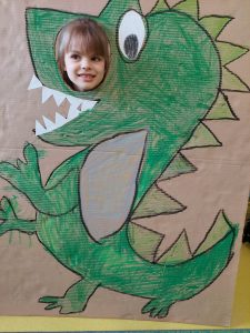 Uczennica pozuje do zdjęcia z makietą przedstawiającą odręcznie namalowanego zielonego dinozaura. Makieta obok oka dinozaura ma okrągły otwór przez, który wystawia głowę uśmiechnięta uczennica.