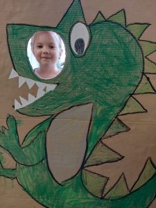 Uczennica pozuje do zdjęcia z makietą przedstawiającą odręcznie namalowanego zielonego dinozaura. Makieta obok oka dinozaura ma okrągły otwór przez, który wystawia głowę uśmiechnięta uczennica.