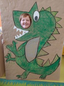 Uczennica pozuje do zdjęcia z makietą przedstawiającą odręcznie namalowanego zielonego dinozaura. Makieta obok oka dinozaura ma okrągły otwór przez, który wystawia głowę uśmiechnięta uczennica.