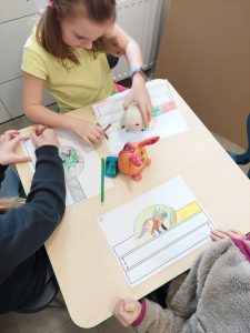 Trzy uczennice siedzą przy stoliku i kolorują leżące przed nimi rysunki. Pośrodku stolika ustawiona jest maskotka.