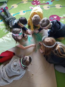 Uczniowie leżą na dywanie. Pośrodku między nimi leży tekturowa makieta z obrysem dinozaura. Dzieci malują dinozaura na zielono.