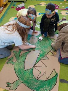 Uczniowie podczas wykonywania makiety z dinozaurem. Dzieci malują na zielono dinozaura i przyklejają mu zęby. Makieta leży na kolorowym dywanie a dzieci kucają wokół niej.