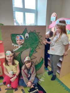 Uczniowie pozują do wspólnego zdjęcia przy makiecie z narysowanym zielonym dinozaurem. Dwie uczennice siedzą przed makietą. Obok makiety stoi dziewczynka, która wraz z kolegą wychylającym się zza makiety, ją podtrzymuje. W makiecie na wysokości oka dinozaura jest okrągły otwór, przez który wystawia głowę kolejny uczeń.