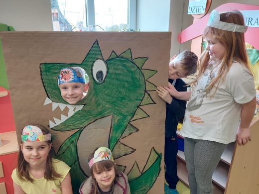 Uczniowie pozują do wspólnego zdjęcia przy makiecie z narysowanym zielonym dinozaurem. Dwie uczennice siedzą przed makietą. Obok makiety stoi dziewczynka, która wraz z kolegą wychylającym się zza makiety, ją podtrzymuje. W makiecie na wysokości oka dinozaura jest okrągły otwór, przez który wystawia głowę kolejny uczeń.